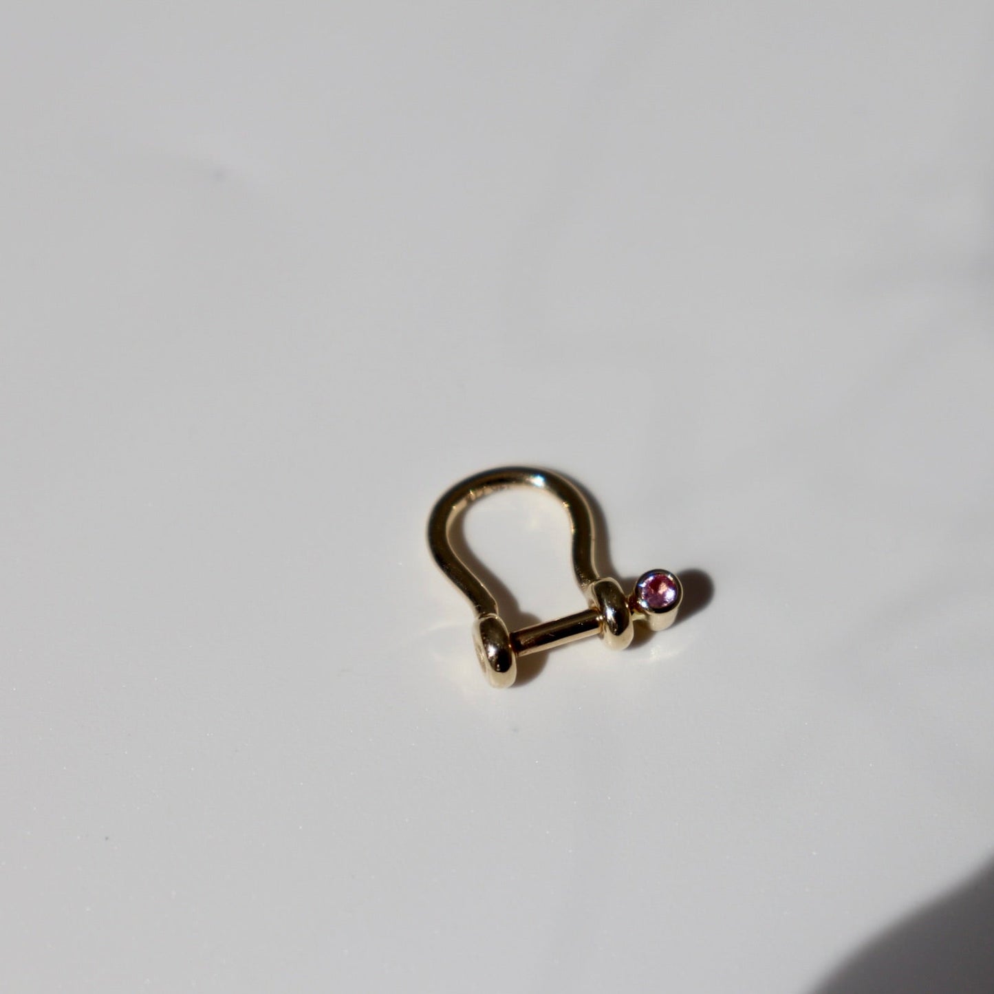 A small gold-toned metal clasp or toggle with a tiny pink gemstone accent, photographed on a white surface with strong sunlight and a dark shadow cast across the right side.