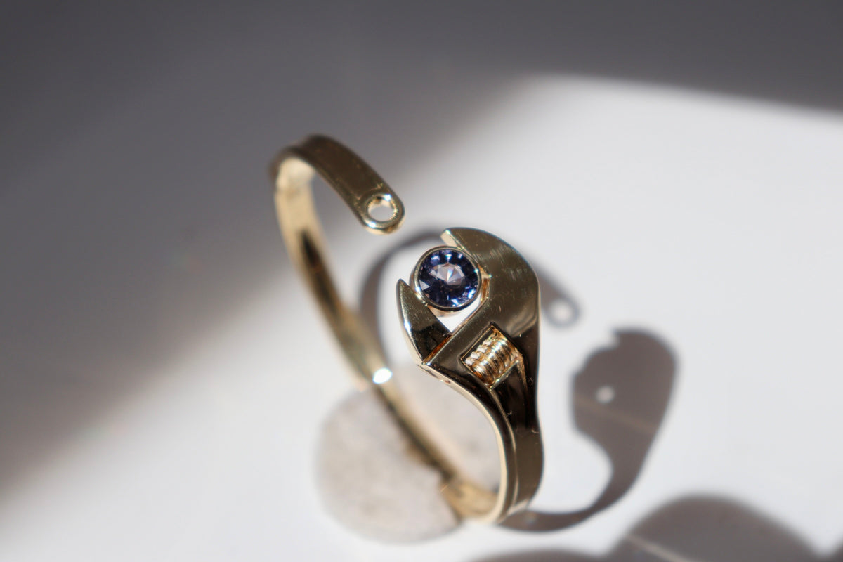 A gold-toned open bangle ring with a central round blue gemstone and a small side accent, photographed on a white surface with strong sunlight and shadows.