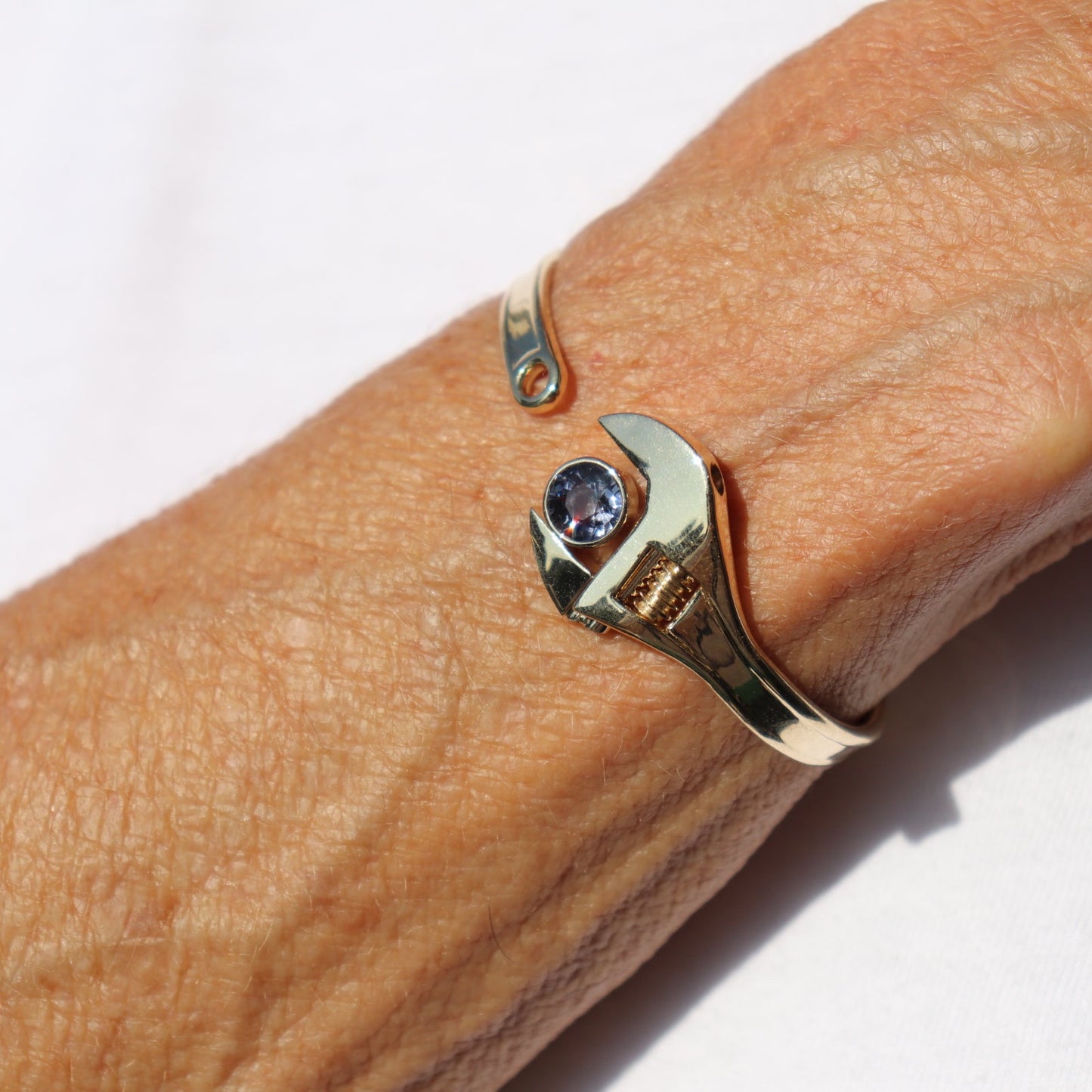 A close-up of a person's wrist wearing a gold wrench-shaped cuff bracelet with a small round purple gemstone in the center, set against a white background.