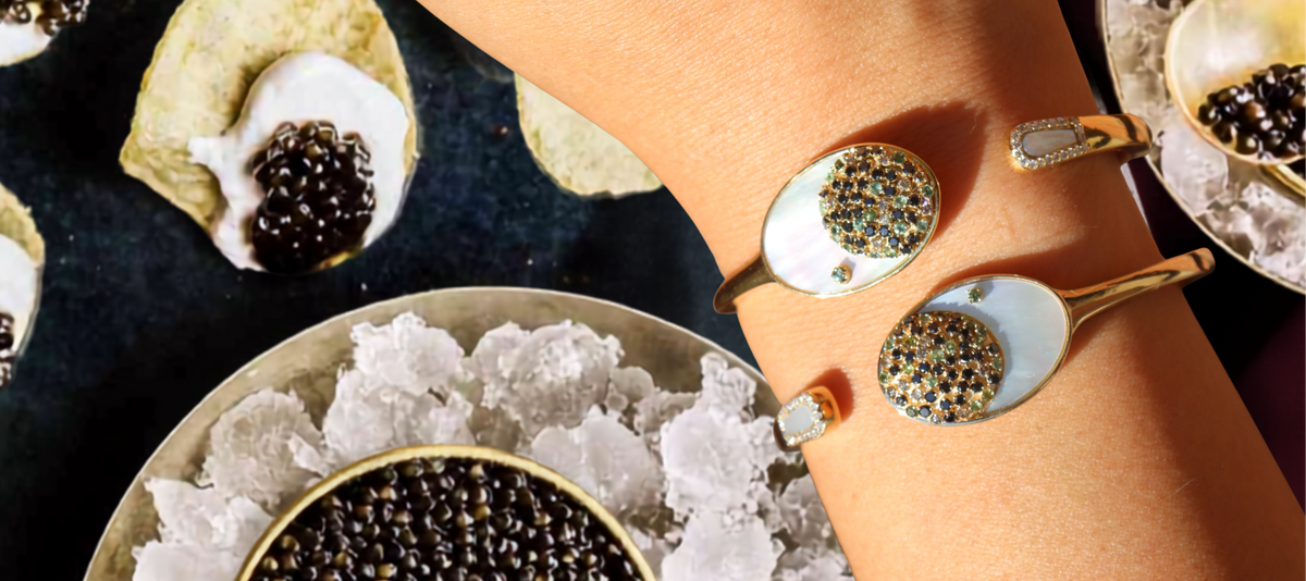 A close-up lifestyle shot of a person's wrist wearing two gold-toned cuff bracelets with white mother-of-pearl spoon inlays and sparkling crystal-encrusted oval centers. The scene is styled with a bowl of black caviar on crushed ice, with additional caviar and chips blurred in the background.